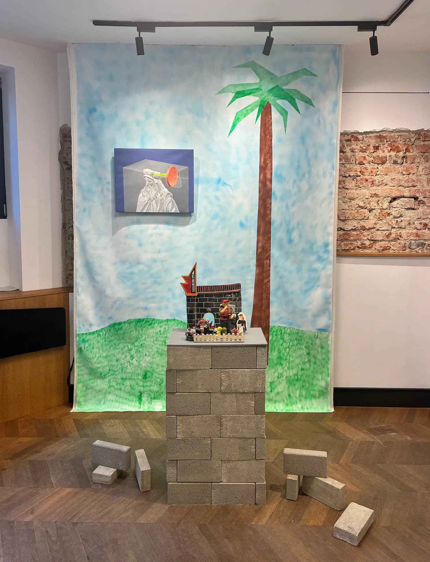 Wide gallery view with the painted backdrop, a small painting, and the brick-block store sculpture on its pedestal