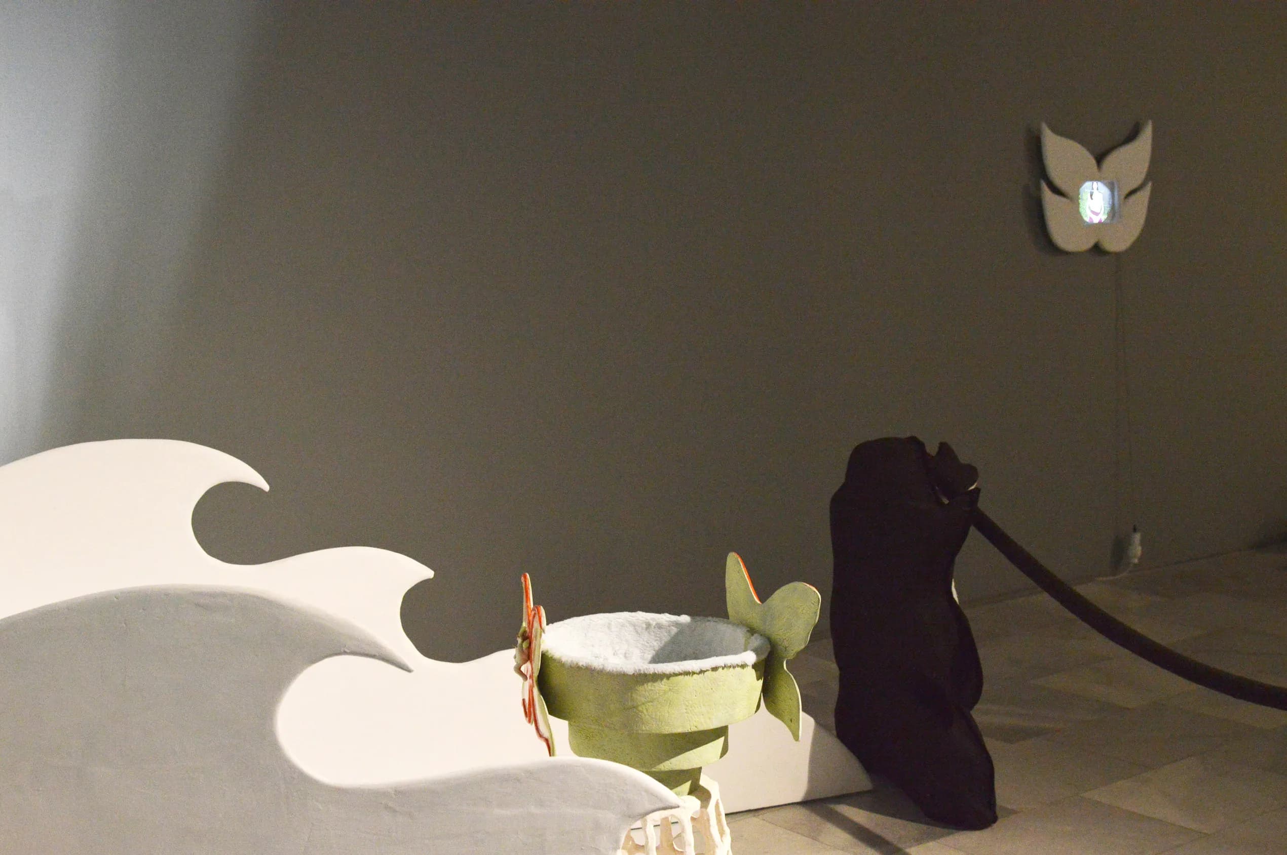 Installation view with white wave-shaped cutouts in the foreground, a ceramic bowl on a plinth, a dark draped figure, and the wall-mounted mask with its glowing screen