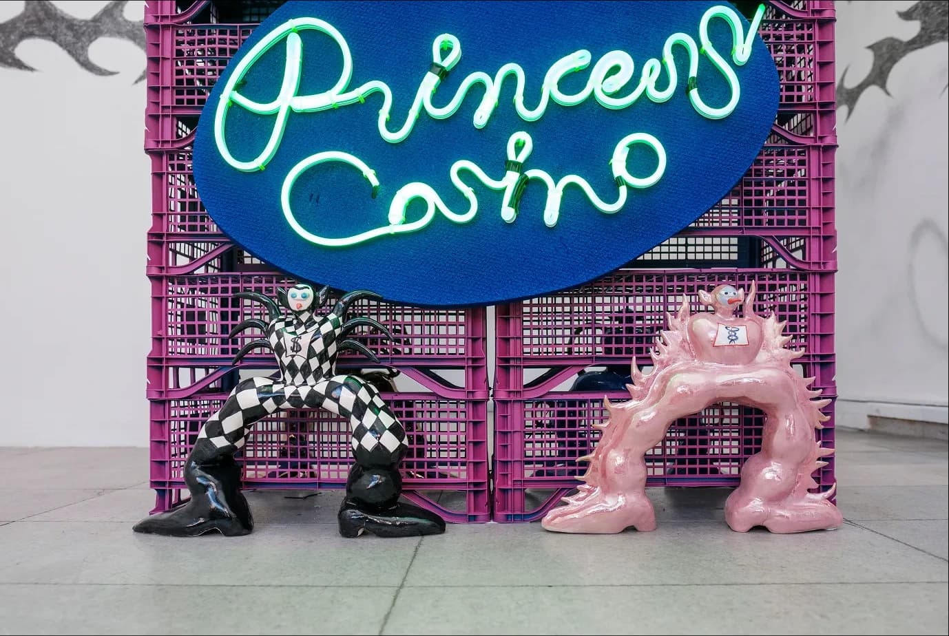 Neon 'Princess Casino' sign above a checkered harlequin figure and a spiked pink figure standing under a crate table