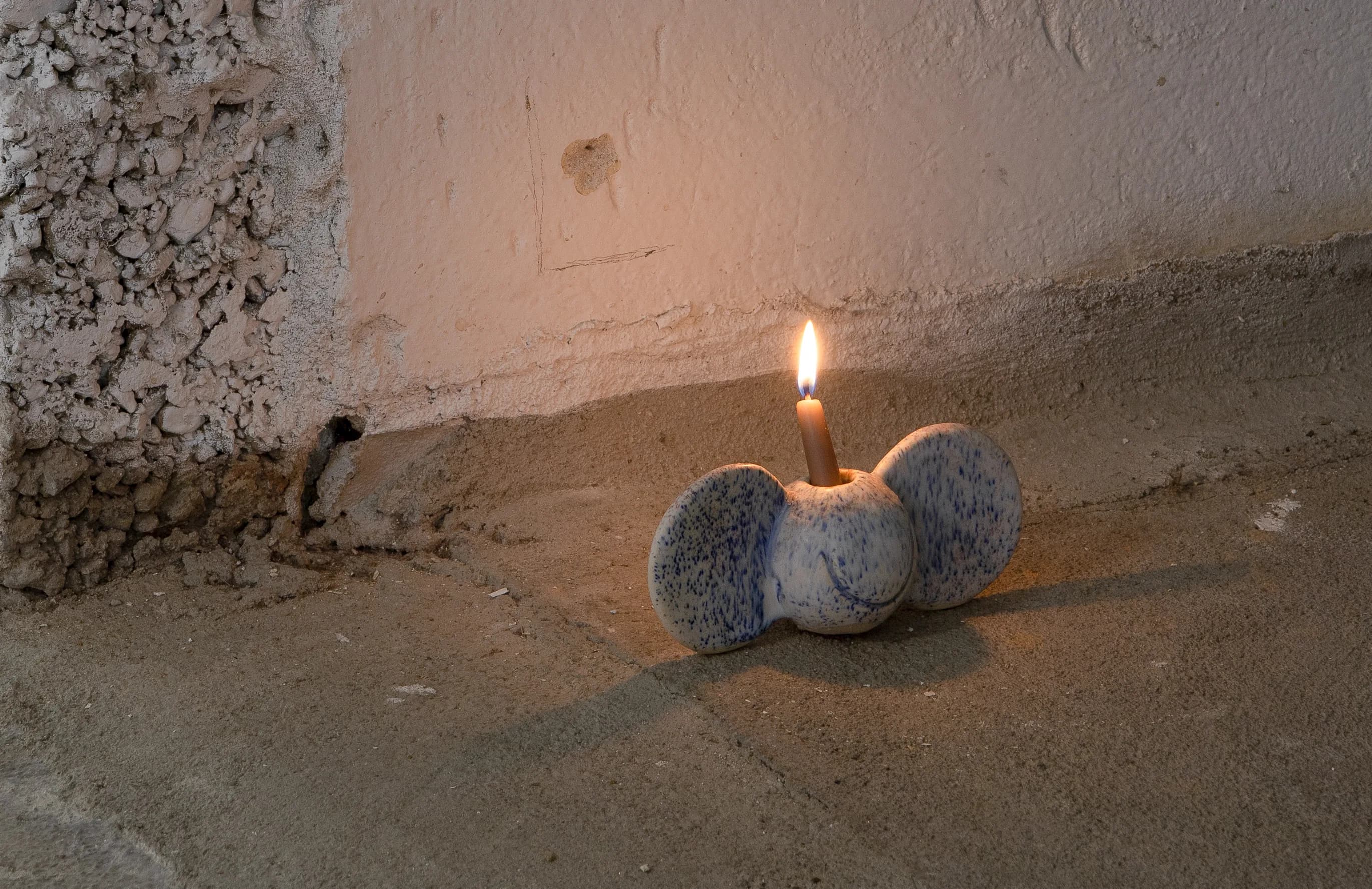 A lit candle in a blue-speckled ceramic candle holder with round ears, standing on the gallery floor